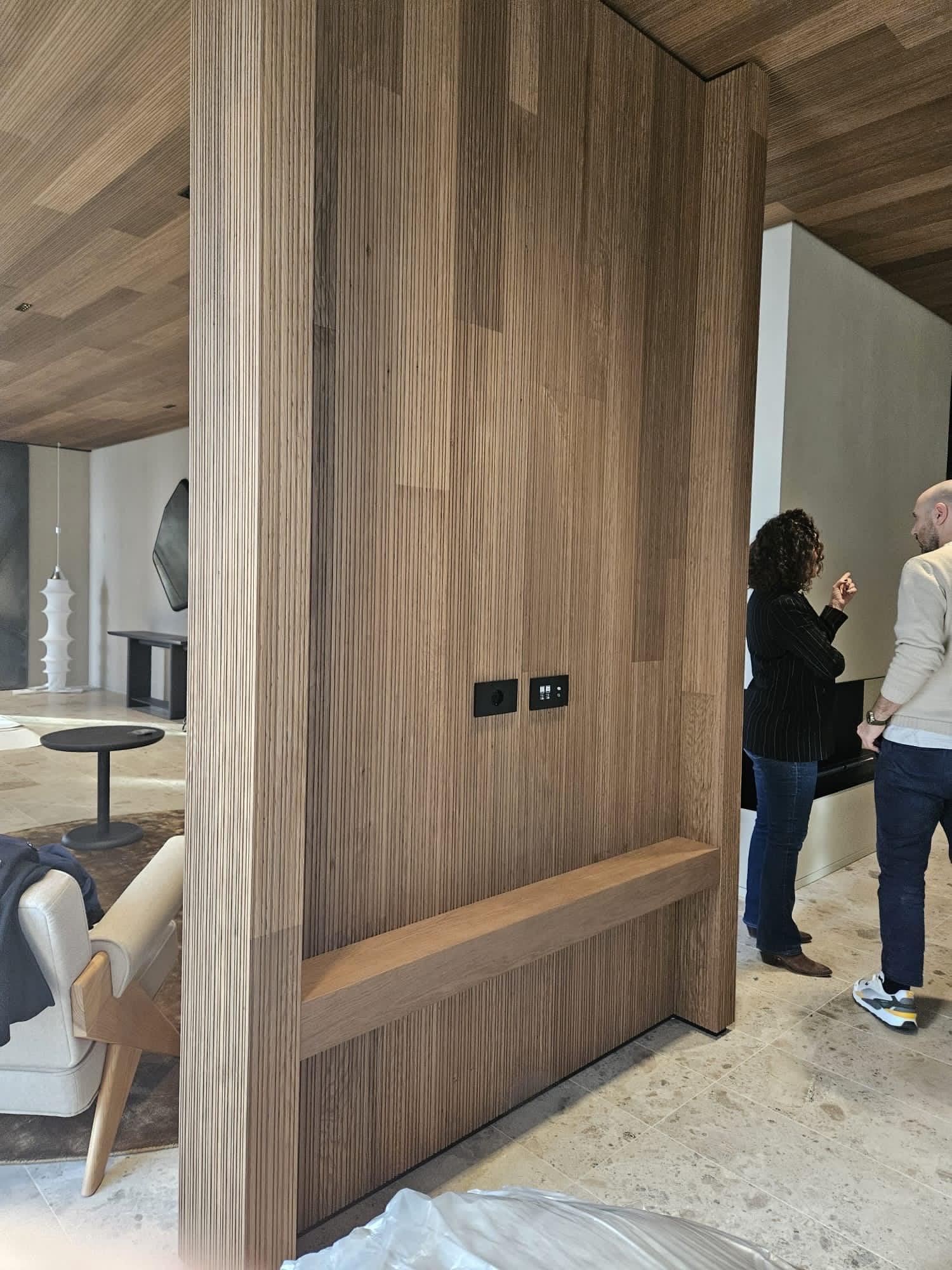 Side of a Slat fluted oak column with a built-in bench ledge and black switch plates, under a matching slatted ceiling