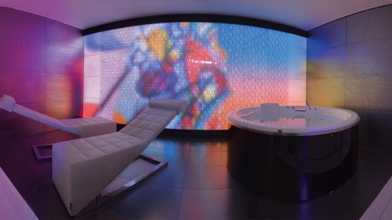 Curved backlit color-changing Luminous panel behind a chaise lounge and soaking tub in a spa treatment room